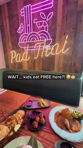 A top-down, flat-lay shot of a table filled with various Asian dishes, including curries, noodles, and starters, set against a wooden wall with a neon 'Pad Thai' sign in the background.