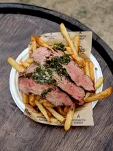 A top-down, close-up shot of a steak and fries dish served on branded paper in a paper plate, resting on a wooden barrel. The steak is sliced and topped with a vibrant green chimichurri sauce, appearing juicy and well-cooked.
