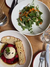 An overhead, natural-light photograph of two plates of food on a wooden table, featuring a burrata salad with strawberries and a smoked mackerel salad.