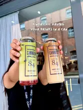 A promotional shot featuring two canned drinks, a matcha and an iced coffee, held up in front of a brick building at a canal-side venue.