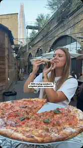 A POV-style shot of a person eating a large pizza at an outdoor venue in London, with The Shard visible in the background.