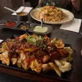 A close-up, slightly dark shot of a sizzling steak and chips platter served in a restaurant, with another dish visible in the background.