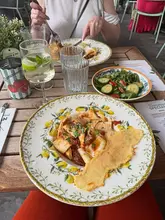 A casual, bright, and inviting photo taken from an overhead perspective at an Ask Italian restaurant. The image features a plate of beef pappardelle, a side salad, and a Hugo Spritz, set against a wooden table with lemon-patterned crockery. The natural lighting and outdoor setting create a relaxed, summery dining atmosphere.