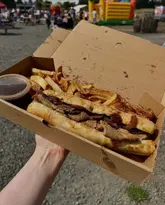A casual, first-person perspective shot of a French dip sandwich and chips in a cardboard takeaway box, held up against a busy outdoor event background featuring a bouncy castle and farm shop setting.