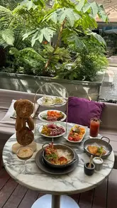 A short, engaging reel showcasing a brunch experience at the Sanderson Hotel in London, featuring a large bagel tower with various toppings, lobster mac and cheese, and desserts in a scenic courtyard setting.