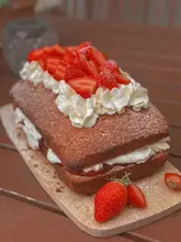 A high-quality, close-up photograph of a homemade Victoria Sponge loaf cake, topped with fresh strawberries and whipped cream, presented on a rustic wooden board.