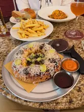 A flat-lay, overhead shot of a restaurant table featuring a round pizza-style dish topped with olives and cheese, a plate of fries, a bowl of pasta, bread, and a glass of orange wine.