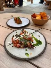 A high-quality, overhead shot of a Greek-Japanese fusion meal served outdoors. The image features a plate of prawn saganaki in the foreground, accompanied by a skewer and a bowl of crispy potatoes, all set on a wooden table. The lighting is natural and bright, highlighting the textures and colors of the food.