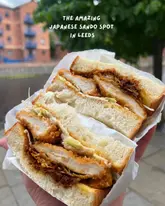 A high-quality, close-up shot of a chicken katsu sandwich held in hand, effectively highlighting the texture of the fluffy milk bread and the crispy coating of the chicken. The shallow depth of field keeps the focus entirely on the food, making it look fresh and highly desirable.