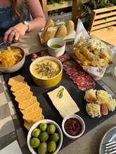 A high-quality, overhead flat-lay shot of a charcuterie and cheese board, accompanied by truffle chips and wine, set on an outdoor table. The lighting is bright and natural, highlighting the textures of the food.