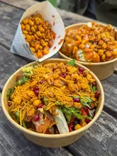 A close-up shot of Indian street food served in paper bowls and a cone, placed on a rustic wooden table. The image features a samosa chaat, chicken tikka popcorn, and jhalmuri corn, showcasing vibrant colours and textures typical of Indian street food.