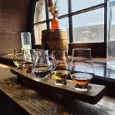 A well-composed, atmospheric shot of a whisky tasting flight served on a wooden board, positioned on a window ledge in a rustic bar setting.