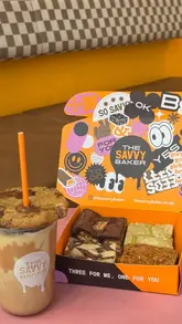 A short, energetic video showcasing a visit to The Savvy Baker in Leeds, highlighting their cookie butter iced latte topped with a cookie and a selection of brownies.