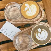 A clean, professional flat lay photograph of two specialty coffees served on wooden trays, accompanied by a menu card detailing the coffee's origin and tasting notes. The composition is balanced and highlights the latte art effectively.