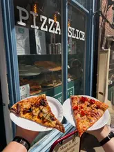A casual, first-person shot of two slices of pizza held up in front of a shop window, showcasing a casual street food experience in Lincoln.