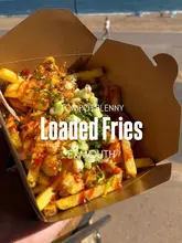 A simple, effective shot of loaded fries held in front of a beach backdrop, clearly communicating the casual, seaside nature of the venue. The lighting is natural and bright, highlighting the textures of the food, while the background provides context without distracting from the main subject.