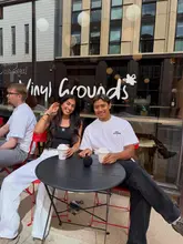 A lifestyle shot of two people sitting at an outdoor table at Vinyl Grounds, holding coffee cups, with the venue's branding visible on the window behind them.