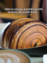 A high-quality, close-up shot of a chocolate-striped pastry, likely a pain au chocolat, served in a paper tray. The image uses a shallow depth of field to blur a coffee cup in the foreground, creating a professional, inviting aesthetic typical of high-end bakery marketing.
