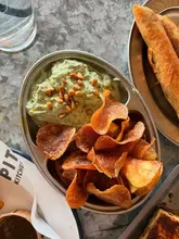 A close-up, top-down shot of a metal bowl filled with homemade crisps and a side of tzatziki dip, served on a metal table.