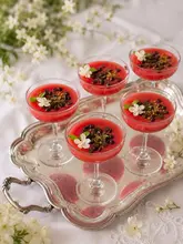 A beautifully styled, high-quality photograph of Gelo di Melone, a traditional Sicilian watermelon pudding, served in elegant coupe glasses on a silver tray. The dessert is garnished with dark chocolate chips, crushed pistachios, and delicate white jasmine flowers, set against a soft, bright background.