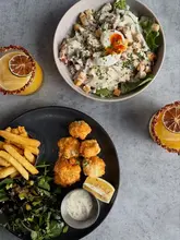 A high-quality, overhead flat-lay photograph of a lunch or dinner spread featuring a chicken caesar salad, a plate of monkfish scampi with fries, and two mango chilli margaritas. The setting is clean and modern, focusing entirely on the food and drink presentation.