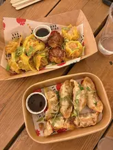 A top-down, overhead shot of two takeaway containers filled with dumplings and gyoza, served with dipping sauce, placed on a wooden table.