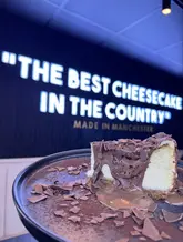 A close-up shot of a chocolate cheesecake slice, positioned in front of a neon sign that reads 'The Best Cheesecake In The Country'. The image is intended to showcase the indulgent nature of the dessert at a new venue.