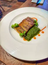 A high-quality, well-composed food photograph of a pressed lamb starter served at The Bear in Tattenhall. The image captures the dish from an appropriate angle, highlighting the texture of the meat and the vibrant green chimichurri, set against a clean, rustic wooden table background.