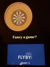 A promotional image for an interactive darts venue, featuring a lit dartboard and a digital scoring screen, designed to highlight the activity-based social experience.