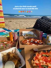 A casual, outdoor beach picnic scene featuring takeaway pizza, fried chicken, and canned cocktails. The image captures a relaxed, sunny vibe, though the presentation is purely functional rather than styled.