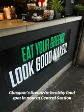A promotional image for a healthy food venue located in Glasgow Central Station, featuring a prominent sign with the text 'Eat your greens, look good naked' above a service counter.