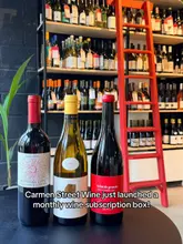 A promotional image for a wine subscription box featuring three bottles of wine arranged on a table in front of a wine shelf.