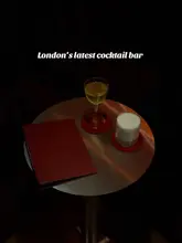 A high-quality, moody promotional shot of two cocktails and a menu on a small table in a dimly lit London bar. The image focuses on the presentation of the drinks, creating an intimate and sophisticated atmosphere.