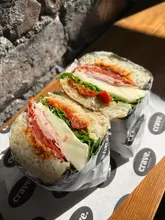 A close-up, high-quality photograph of a 'Godfather' sandwich served on branded paper, showcasing the layers of meat, cheese, and sun-dried tomatoes within focaccia bread. The natural lighting highlights the textures of the ingredients, making the food look fresh and appealing.