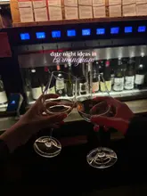 A first-person perspective shot of two wine glasses being toasted in front of a wine dispensing machine, setting a romantic and atmospheric tone for a date night.