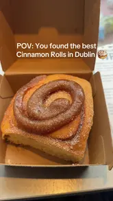 A short, handheld vlog-style video featuring a visit to a cafe in Dublin. The creator pans across the display case, highlighting various cinnamon rolls and cookies, before taking a bite of a cinnamon roll to show the texture.