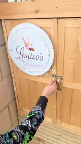 A mother and her young son visit a local 'Cake Shed' to pick up some Easter treats. The video shows the exterior of the small, self-service wooden shed, the interior shelves stocked with various packaged baked goods like fudge, brownies, and cheesecake boxes, and the final haul of items they purchased.