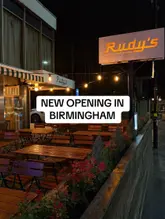 A night-time exterior shot of a newly opened Rudy's Pizza Napoletana branch, featuring outdoor seating and illuminated signage.