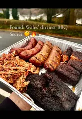 A food review video featuring a takeaway tray of Texas-style BBQ, including pulled pork, beef cheek, sausages, mac and cheese, and beans, filmed by a creator in a car.