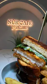 A montage video showcasing the interior and menu items at Nur Café, including drinks, sandwiches, and dessert domes. The video highlights the cafe's aesthetic, food presentation, and amenities like the prayer room.