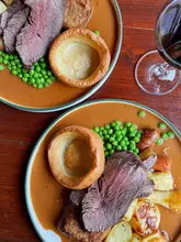 A top-down shot of two Sunday roast beef dinners served on a wooden table, accompanied by a glass of red wine. The plates feature roast beef, Yorkshire puddings, roast potatoes, carrots, and peas, all swimming in a generous amount of stout gravy.