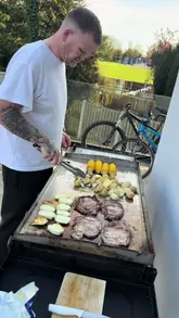 A man prepares a meal of steak, corn on the cob, potatoes, and halloumi on an outdoor flat-top grill in a garden setting.