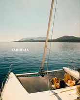 A high-quality, aspirational travel shot taken from a high angle, likely a drone, showing two people relaxing on the netting of a catamaran in the middle of the sea in Sardinia. The composition effectively emphasizes the vastness of the ocean and the relaxing nature of the experience, creating a strong sense of wanderlust.