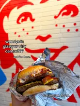 A close-up, first-person shot of a Baconator burger held in front of a branded Wendy's wall, promoting the opening of a new location in Glasgow.