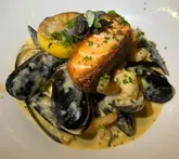 A close-up, top-down shot of a seafood dish featuring a seared salmon fillet, mussels, and shellfish in a creamy bisque sauce, served on a white plate.