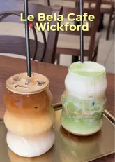 A simple, direct shot of two iced drinks—an iced coffee and an iced matcha latte—served in trendy, wavy-shaped glasses on a gold tray at Le Bela Cafe in Wickford. The image is clearly intended to showcase the cafe's aesthetic drink presentation.