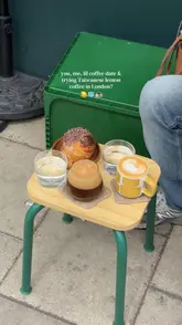 A high-angle, aesthetic shot of a coffee date setup featuring a lemon coffee and a pastry served on a small wooden stool outdoors. The image captures a casual, trendy vibe typical of independent London coffee shops, utilizing natural daylight and a shallow depth of field to focus on the drinks and food.