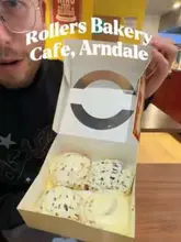 A creator visits Rollers Bakery Cafe in the Arndale Centre, Manchester, to review their cinnamon buns and pizza. The video features the creator talking to the camera, showing the food display, the preparation of the food, and tasting the items.