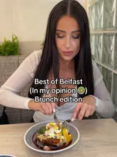 A social media video still featuring a creator reviewing a brunch dish in a restaurant setting. The shot captures the creator mid-bite, with text overlay introducing the content as a 'Best of Belfast' brunch review.