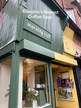 A clear, eye-level photograph of the storefront of 'morning rush' in Glasgow, featuring a distinctive green facade and an adjacent cafe with event posters visible in the window.